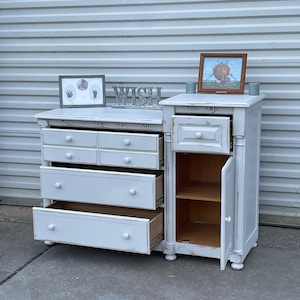 May include: A distressed white dresser and nightstand set with multiple drawers and a cabinet. The dresser drawers are partially open, revealing the interior. Decorative framed artwork and the word "WISH" are on top.