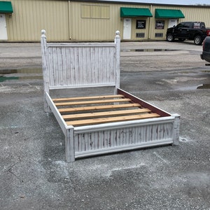 May include: White wooden bed frame with a slatted headboard and footboard. The frame has a distressed white finish and is sitting on a concrete surface.