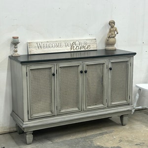 May include: A gray painted wooden cabinet with four doors and a black countertop. The doors have a wire mesh insert and black knobs. The cabinet is sitting on a concrete floor. A sign above the cabinet reads "Welcome to our home".