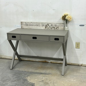 May include: A gray wooden desk with three drawers and a criss-cross base. The desk has a sign on top that says "Welcome to our home".