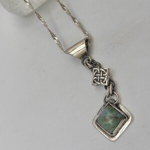May include: A silver necklace with a twisted chain and a pendant featuring a square knot design and a diamond-shaped, green-hued stone. The pendant is framed in a silver setting, creating a vintage aesthetic.