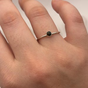 May include: A delicate silver ring featuring a small, round, dark gemstone. The ring's band is thin and simple, highlighting the gemstone's color and sparkle. The ring is shown on a finger.