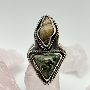 May include: A silver ring with a triangular green stone and a shell-shaped stone. The ring has a rope-like border around the stones and a wide band. The ring is set against a white background with pink stones.