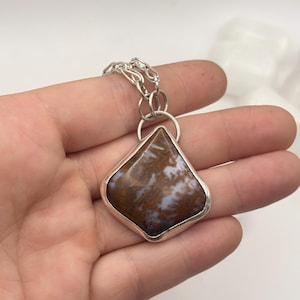 Op de afbeelding: Een zilveren hangerketting met een ruitvormige steen met bruine en witte wervelingen. De steen is gezet in een zilveren rand en hangt aan een zilveren ketting. Deze ketting is een sieraad.