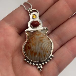 May include: A silver pendant featuring a natural seashell with brown and white radial patterns, set in a decorative silver frame. The pendant includes a small oval red stone and a tiny yellow stone at the top, suspended from a silver chain.