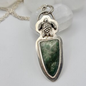 May include: A silver pendant necklace featuring a shield-shaped green stone. A detailed silver turtle is at the top of the pendant. The necklace has a delicate silver chain. The pendant is approximately 2 inches long.