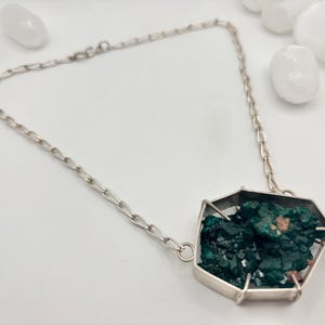 Könnte beinhalten: Silberfarbene Halskette mit einem geometrischen Anhänger mit einem Cluster aus dunkelgrünen Kristallen. Die Halskette hat eine filigrane Kette und liegt vor einem weißen Hintergrund mit weißen Steinen.