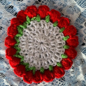 May include: A white crocheted coaster with a red and green crocheted flower border.