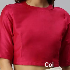 Pode incluir: Uma blusa magenta de manga curta com gola alta. A blusa tem um design simples e elegante e é feita de um tecido liso, possivelmente acetinado. A palavra "Coi" está impressa na parte inferior da blusa.