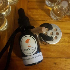 May include: A black glass bottle with a dropper, a label with the text "BAYEB" and "Black Raspberry & Vanilla". A silver metal container with a wooden stick is attached to the bottle. The background is a wooden surface.
