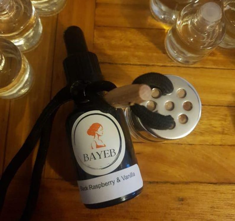 May include: A black glass bottle with a dropper, a label with the text "BAYEB" and "Black Raspberry & Vanilla". A silver metal container with a wooden stick is attached to the bottle. The background is a wooden surface.