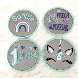Könnte beinhalten: Vier runde, graue Filzpatches mit türkisfarbenem Rand. Die Patches zeigen gestickte Designs: einen Regenbogen mit dem Namen "Sophie", ein Einhorn-Gesicht, ein Banner mit der Zahl "1" und dem Wort "Klasse" sowie eine Krone mit dem Text "Sei Frech Wild Und Wunderbar".