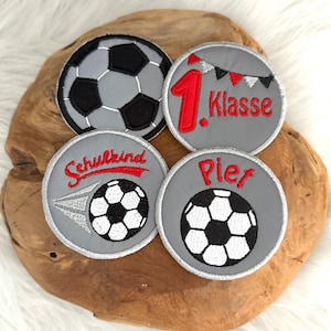 Könnte beinhalten: Vier graue und schwarze Fußballaufnäher mit gestickten Details. Einer trägt die Aufschrift "1. Klasse" in Rot, ein anderer "Schulkind" und ein dritter "Pier". Die Aufnäher liegen auf einer Holzoberfläche.