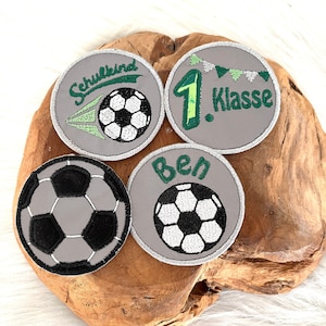Könnte beinhalten: Vier graue Filz-Bügeletiketten mit grüner und weißer Stickerei. Ein Etikett sagt "Schulkid" mit einem Fußball und grün-weißen Streifen. Ein weiteres Etikett sagt "1. Klasse" mit einer grünen Eins und einer Reihe grün-weißer Flaggen. Das dritte Etikett sagt "Ben" mit einem Fußball. Das vierte Etikett ist ein Fußball.