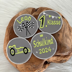 Könnte beinhalten: Vier graue, gestickte Bügelbilder mit gelben und weißen Akzenten. Die Patches zeigen ein Rennwagen, eine karierte Flagge, den Text "Leon", die Zahl "1" mit dem Text "Klasse" und den Text "Schulkind 2024".