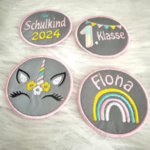 Könnte beinhalten: Vier graue Filzpatches mit rosa Nähten. Ein Patch sagt "Schulkind 2024" mit einer Krone über dem Text. Ein weiterer Patch sagt "1. Klasse" mit einer Flaggenkette über der Zahl. Ein dritter Patch hat ein Einhorn mit einem Regenbogenhorn und Blumen. Der letzte Patch sagt "Fiona" mit einem Regenbogen unter dem Namen.