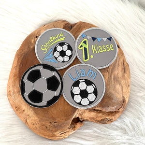 Könnte beinhalten: Vier graue, gestickte Bügelbilder mit einem Fußball-Motiv. Ein Patch sagt "Schulkind" mit einem Fußball und einem gelben und blauen Streifen. Ein weiterer Patch sagt "1. Klasse" mit einem Fußball und blauer und weißer Girlande. Der dritte Patch sagt "Liam" mit einem Fußball. Der vierte Patch hat einen Fußball.