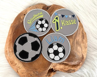 May include: Four gray embroidered iron-on patches with a soccer ball design. One patch says "Schulkind" with a soccer ball and a yellow and blue streak. Another patch says "1. Klasse" with a soccer ball and blue and white bunting. The third patch says "Liam" with a soccer ball. The fourth patch has a soccer ball.