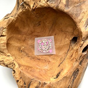 Könnte beinhalten: Ein rosafarbener und brauner Smiley-Aufkleber mit einem Blitz und einem Stern auf einem Holzgrund.
