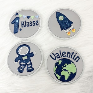 Könnte beinhalten: Vier bestickte Aufnäher mit Weltraum-Motiven. Ein Aufnäher zeigt "1. Klasse" mit der Zahl 1 und einem Banner. Weitere Aufnäher zeigen eine Rakete, einen Astronauten und einen Globus mit dem Namen "Valentin". Alle Aufnäher sind kreisförmig und grau.