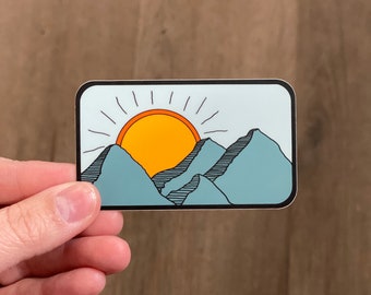 May include: A sticker with a blue, orange, and yellow illustration of a sun rising over mountains. The sticker has a white background and a black border.