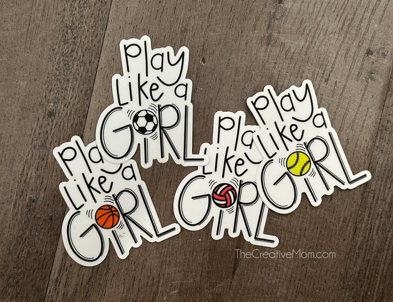 May include: Three white stickers with black text that reads "Play Like a Girl" with a different sports ball on each sticker. The sports balls are a soccer ball, a basketball, and a softball.