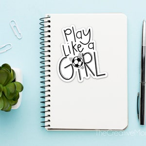 May include: A white notebook with a black and white sticker that says "Play Like a Girl" with a soccer ball graphic. The notebook is on a light blue surface with a small potted plant, two paper clips, and a pen.