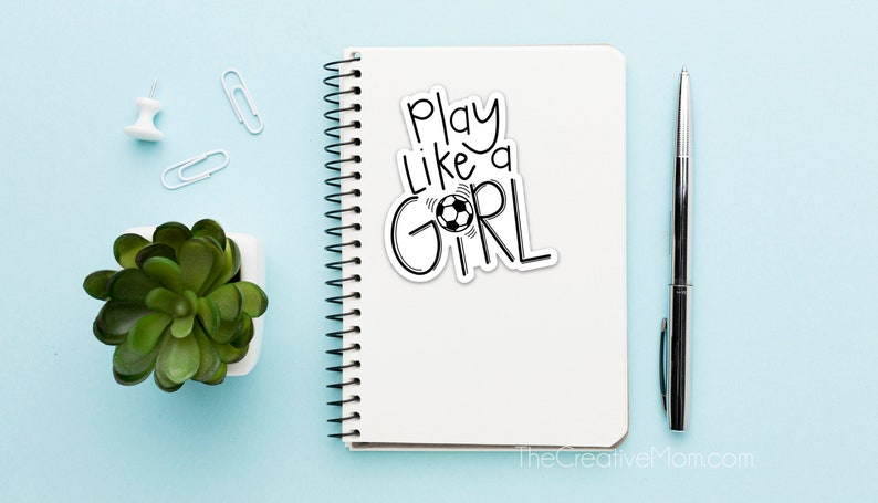 May include: A white notebook with a black and white sticker that says "Play Like a Girl" with a soccer ball graphic. The notebook is on a light blue surface with a small potted plant, two paper clips, and a pen.