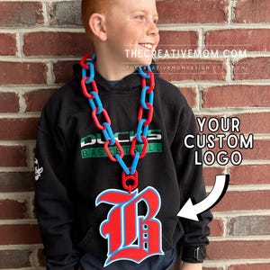 May include: A large, custom-made necklace featuring a thick chain of alternating red and blue plastic links. A prominent, oversized pendant in the shape of a stylized letter B with red, white, and blue coloring hangs from the center of the chain.