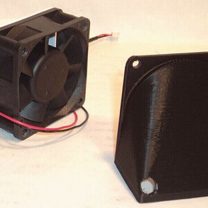 May include: A black computer fan with a white housing and a separate black angled component. The fan has a black impeller and red and black wires. The angled component has mounting holes.