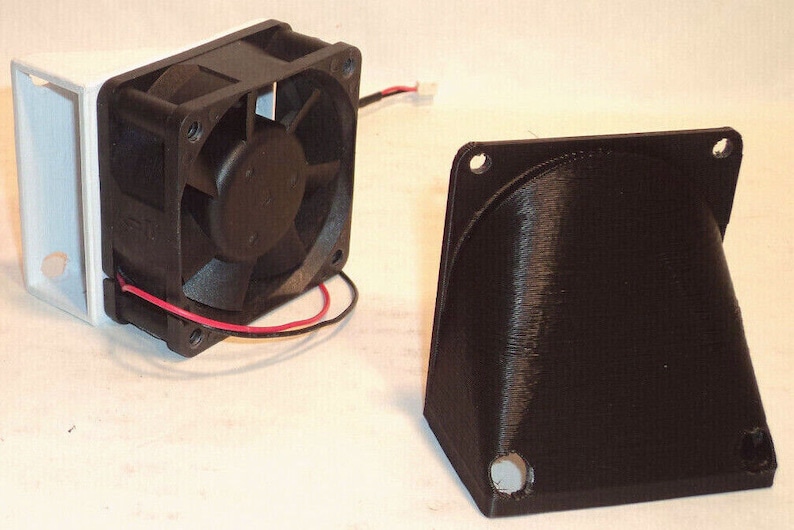 May include: A black computer fan with a white housing and a separate black angled component. The fan has a black impeller and red and black wires. The angled component has mounting holes.