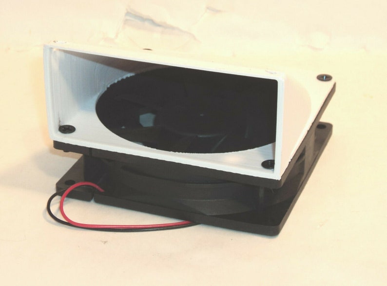 May include: A white plastic fan shroud with a black fan and wires attached. The fan is mounted on a black base.