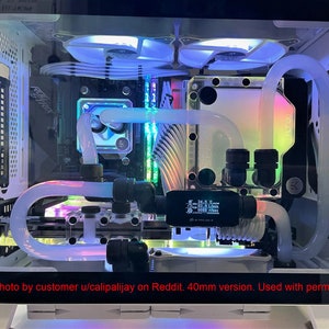 Puede incluir: Una caja de ordenador blanca con un panel lateral transparente, que muestra un circuito de refrigeraci&oacute;n l&iacute;quida personalizado con iluminaci&oacute;n RGB. El circuito incluye un radiador, una bomba, un dep&oacute;sito y tuber&iacute;as. La caja tambi&eacute;n cuenta con una placa base blanca, una tarjeta gr&aacute;fica y otros componentes. El texto "Photo by customer u/calipalijay on Reddit. 40mm version. Used with permission" es visible en la parte inferior de la imagen.