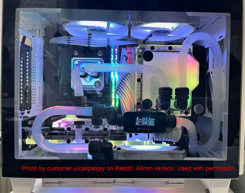 Puede incluir: Una caja de ordenador blanca con un panel lateral transparente, que muestra un circuito de refrigeraci&oacute;n l&iacute;quida personalizado con iluminaci&oacute;n RGB. El circuito incluye un radiador, una bomba, un dep&oacute;sito y tuber&iacute;as. La caja tambi&eacute;n cuenta con una placa base blanca, una tarjeta gr&aacute;fica y otros componentes. El texto "Photo by customer u/calipalijay on Reddit. 40mm version. Used with permission" es visible en la parte inferior de la imagen.