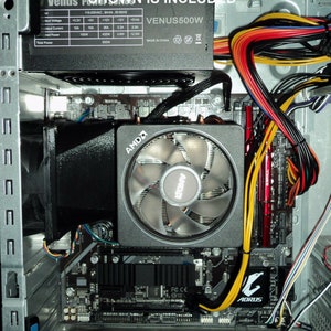 May include: A close-up view of the inside of a computer case. The motherboard is visible, along with a black fan duct, a power supply unit, and various cables. The power supply unit is labeled "VENUS500W" and has a label with specifications. The motherboard has an AMD logo and an AORUS logo.
