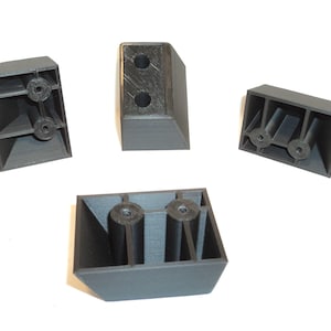 May include: Four dark gray plastic furniture feet. Each foot has a rectangular shape with a beveled edge and a recessed area for mounting. The feet are designed to provide support and stability for furniture.