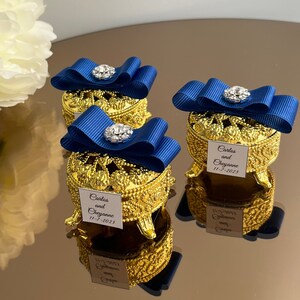 May include: Gold-coloured trinket boxes with dark blue ribbon bows and crystal embellishments. Each box has a tag with the names "Carlos and Cheyenne" and the date "11-7-2023". The boxes are presented on a reflective surface.