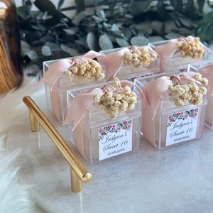 May include: A white marble tray with a gold handle, holding clear boxes with pink ribbon and dried flowers. Each box has a label that says "Jastynn's Sweet 16" with the date "10-06-2023".