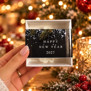 May include: A square white gift box containing a white object, wrapped in a black paper band featuring a graphic of green pine needles and white snow. The text on the band reads HAPPY NEW YEAR 2027 in white capital letters with a serif font.