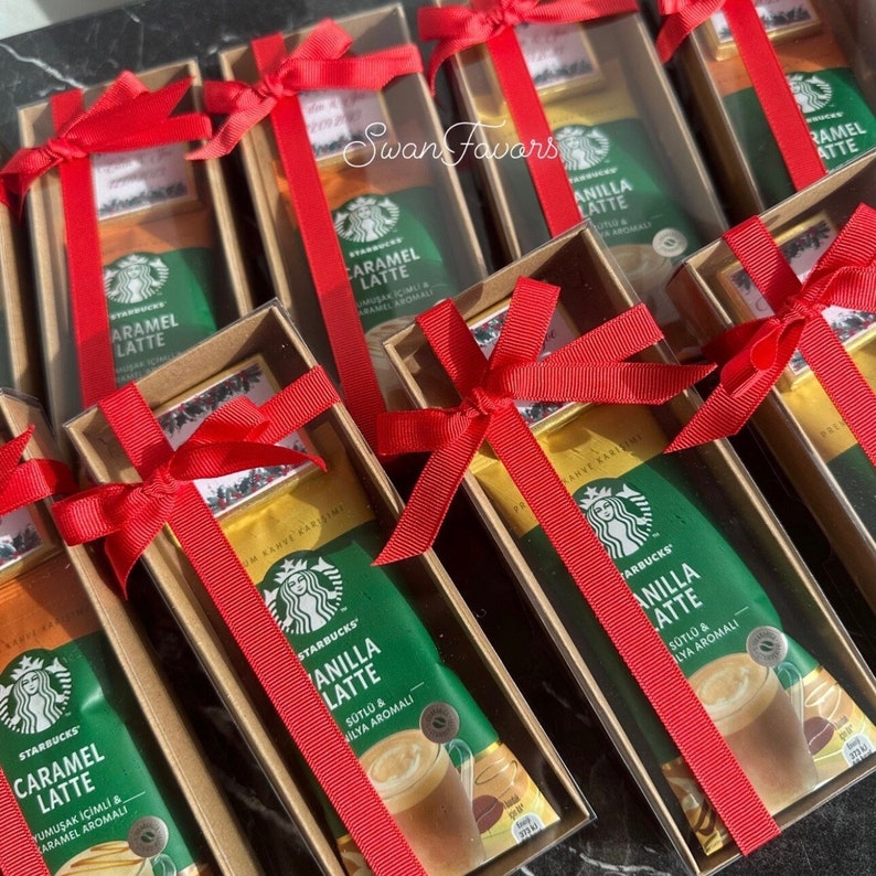 May include: Multiple rectangular cardboard gift boxes containing Starbucks instant coffee packets in Caramel Latte and Vanilla Latte flavors. Each box is tied with a bright red ribbon and includes a small decorative tag with holiday imagery.