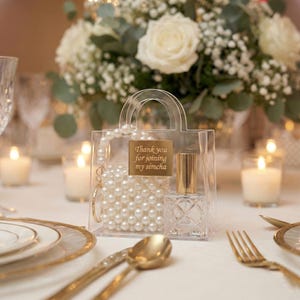 May include: A small, clear handbag filled with faux pearls and a gold-topped perfume bottle. The bag has a gold tag that reads "Thank you for joining my simcha." A gold earring and a pearl necklace are also inside. The bag is on a table with gold cutlery.
