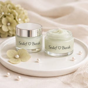 Op de afbeelding: Twee kleine glazen potjes crème met zilveren deksels en de tekst "Sedef ♡ Burak" op de voorkant. De potjes staan op een wit bord met parels en een bloem. De crème binnenin heeft een lichtgroene kleur.