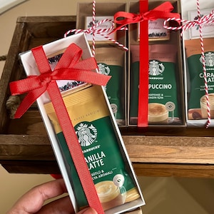 May include: Three rectangular gift boxes containing Starbucks instant coffee packets and small square chocolates. Each box is tied with a bright red ribbon and features a white interior. The coffee packets are green and white with text including Vanilla Latte, Cappuccino, and Caramel Latte.