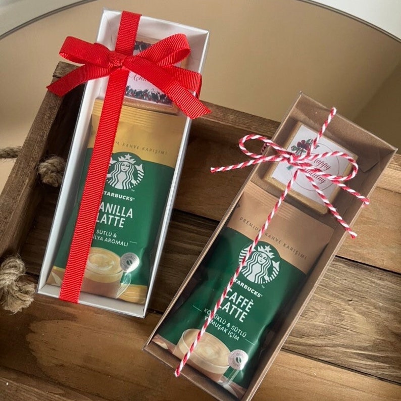 May include: Two rectangular gift boxes containing Starbucks coffee packets and small square cards. The left box is white with a red ribbon, holding a Vanilla Latte packet. The right box is brown with red and white twine, holding a Caffe Latte packet.