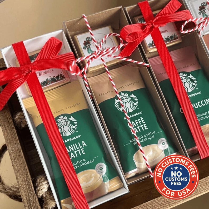 May include: Three rectangular gift boxes containing Starbucks instant coffee packets and small chocolate bars. Each box is tied with a red ribbon and a red and white twine string. The coffee packets are labeled Vanilla Latte and Caffe Latte.