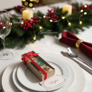 May include: A rectangular cardboard gift box tied with a red ribbon, containing a small item, placed on a white plate. A white place card with a gold border and the name Taylor written in cursive script rests next to the box on the plate.