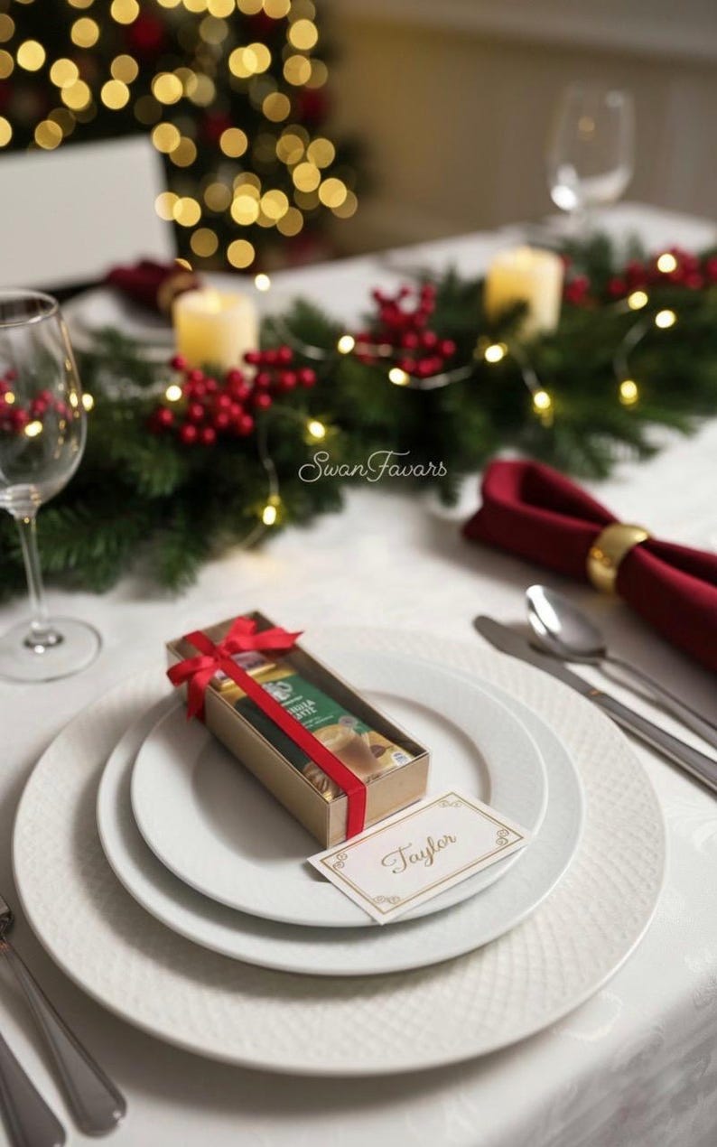 May include: A rectangular cardboard gift box tied with a red ribbon, containing a small item, placed on a white plate. A white place card with a gold border and the name Taylor written in cursive script rests next to the box on the plate.