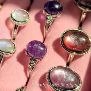 May include: A collection of silver rings with various gemstone settings. The rings feature round, oval, and rectangular stones in shades of purple, pink, and white. The rings are displayed on a pink background.