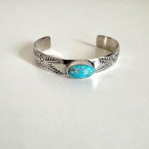 May include: A silver cuff bracelet featuring a central oval turquoise stone with a bright blue hue. The metal band is decorated with etched leaf patterns and circular motifs on both sides of the stone, creating a detailed metallic finish.