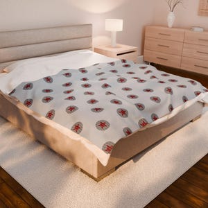 May include: A light-coloured blanket featuring a repeating pattern of red star-like shapes within grey circles. The blanket is laid over a bed with a white sheet and a beige bed frame. The room has a neutral colour palette.
